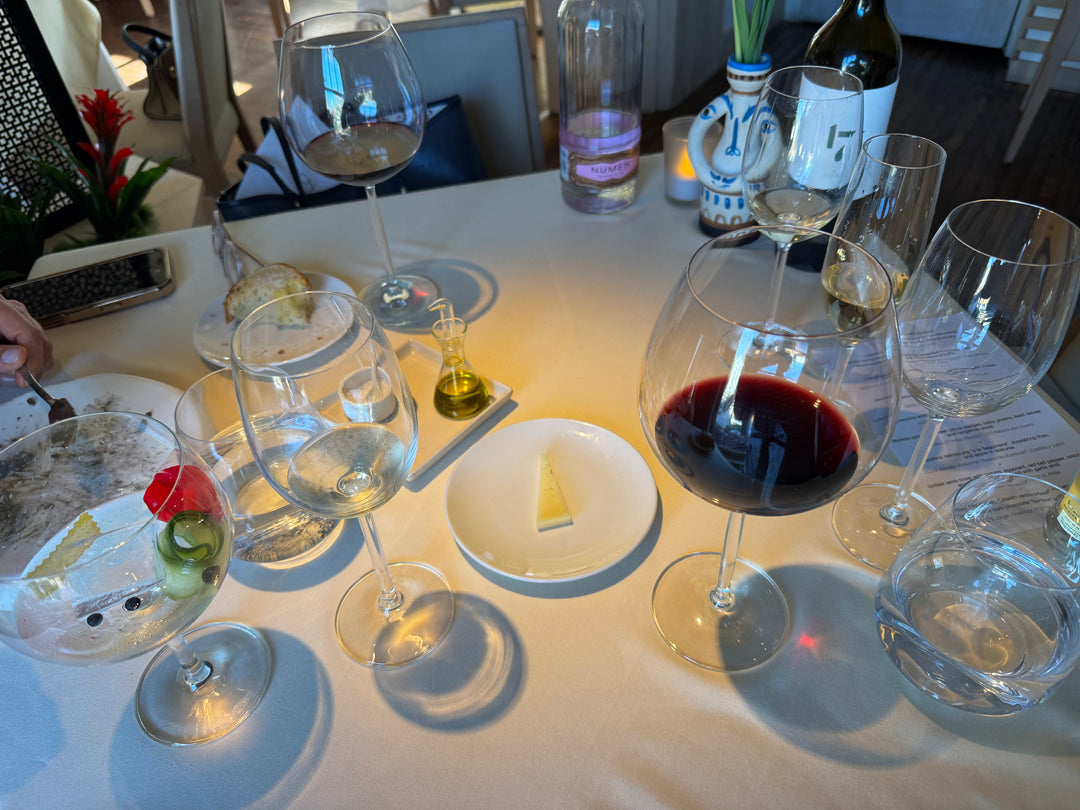 A dining table setting with multiple wine glasses, including one filled with red wine, water glasses, a small bottle of olive oil, and a plate with a butter slice, in a restaurant environment.