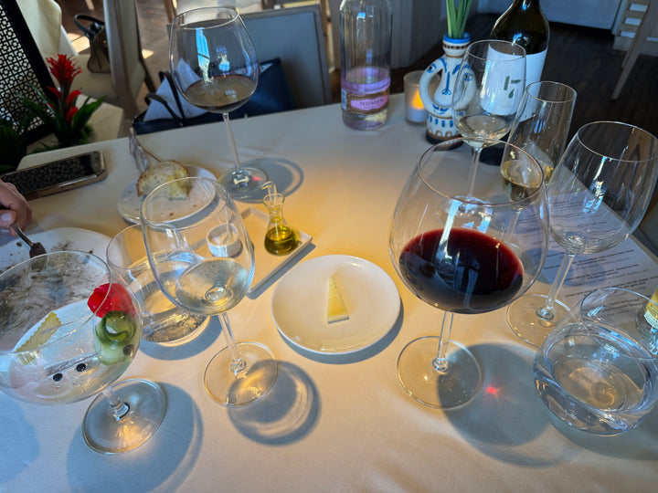 A dining table setting with multiple wine glasses, including one filled with red wine, water glasses, a small bottle of olive oil, and a plate with a butter slice, in a restaurant environment.