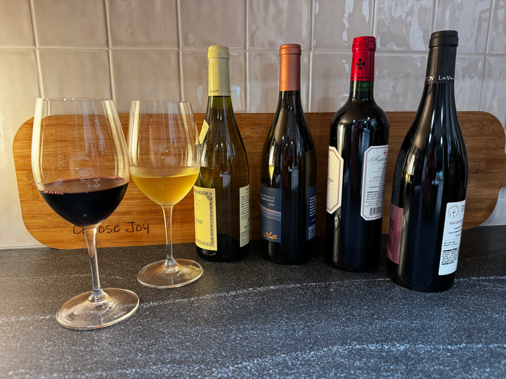 Four wine bottles, reds and whites, from the Munk Mafia Wine Club Mixed bundle on a gray black granite surface with a rustic "Choose Joy" wooden background in warm light.