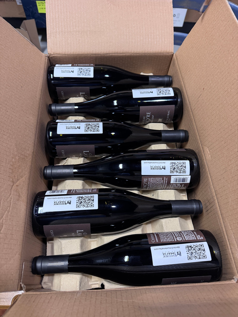 Six red wine bottles with Munk Mafia labels and QR codes arranged inside an open cardboard box on a light paper insert, ready for shipment for a special order.