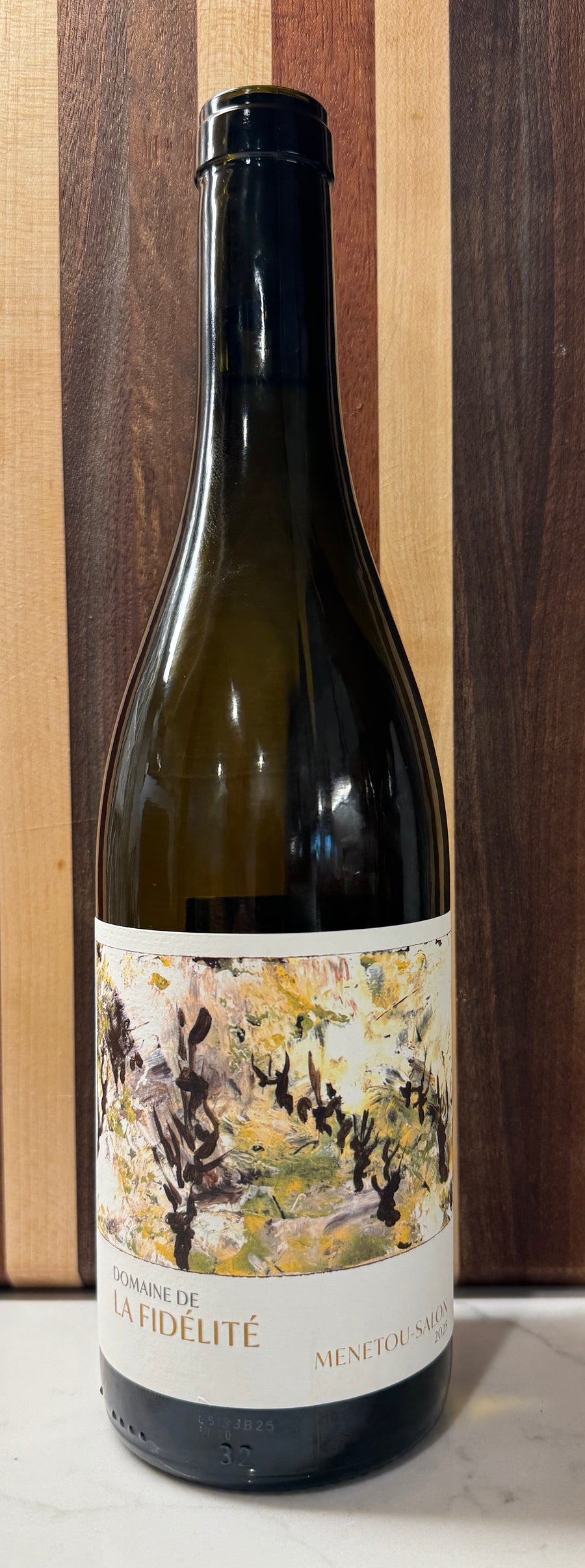 A 2025 Domain de la Fidelite Menetou-Salon Sauvignon Blanc, dark glass wine bottle with a black cap and a cream-colored label featuring an impressionistic painting, set against a vertical wood panel background.