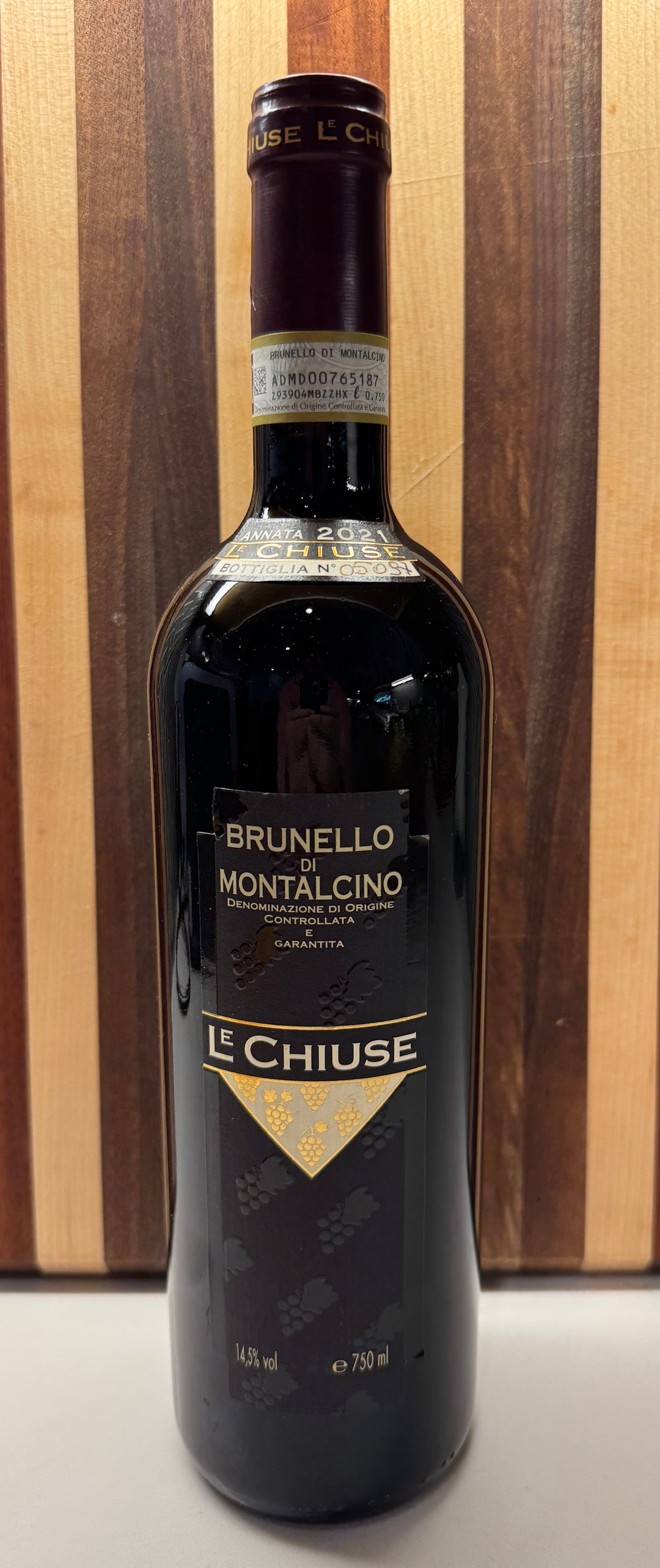 A bottle of Le Chiuse Brunello Montalcino red wine, featuring a dark glass bottle with a gold and black label, set against a wooden striped background.