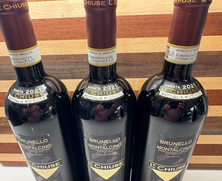 Three bottles of Le Chiuse Annata 2021 Brunello Montalcino bottle No. 05088, 05091, & 05097 red wine, arranged side-by-side against a wooden background.