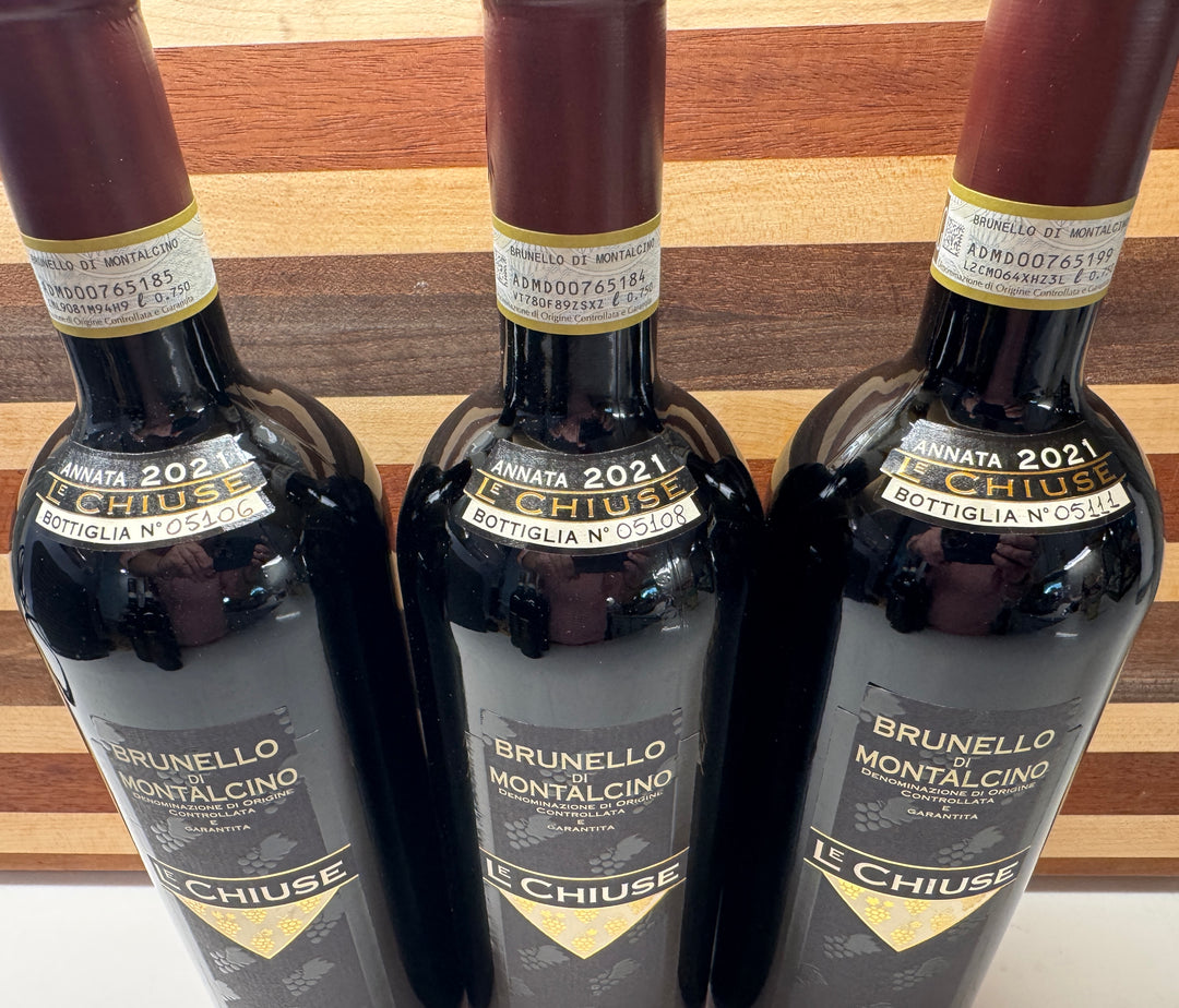 Three dark glass bottles of Le Chiuse Brunello Montalcino bottle No. 05106, 05108, & 05111 red wine lined up against a wooden background.