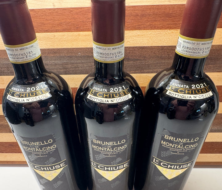 Three dark glass bottles of Le Chiuse Brunello Montalcino bottle No. 05106, 05108, & 05111 red wine lined up against a wooden background.